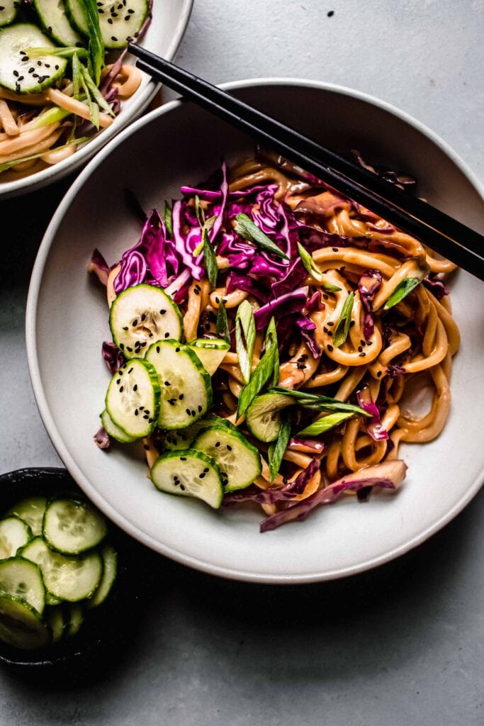 Overhead shot of cold peanut noodle salad in white bowl next to small bowl of pickled cucumbers.