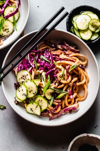 Overhead shot of cold peanut noodle salad in white bowl next to small bowl of pickled cucumbers.