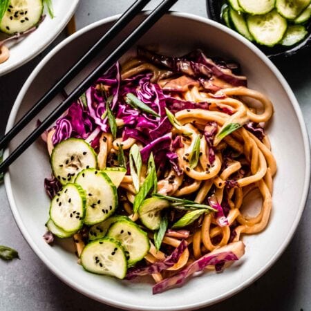 Overhead shot of cold peanut noodle salad in white bowl next to small bowl of pickled cucumbers.