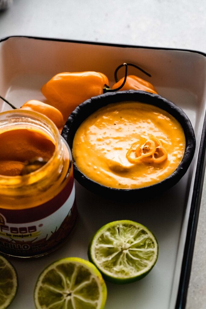 Side view of aji amarillo in black bowl surrounded by limes, peppers, and aji paste. 