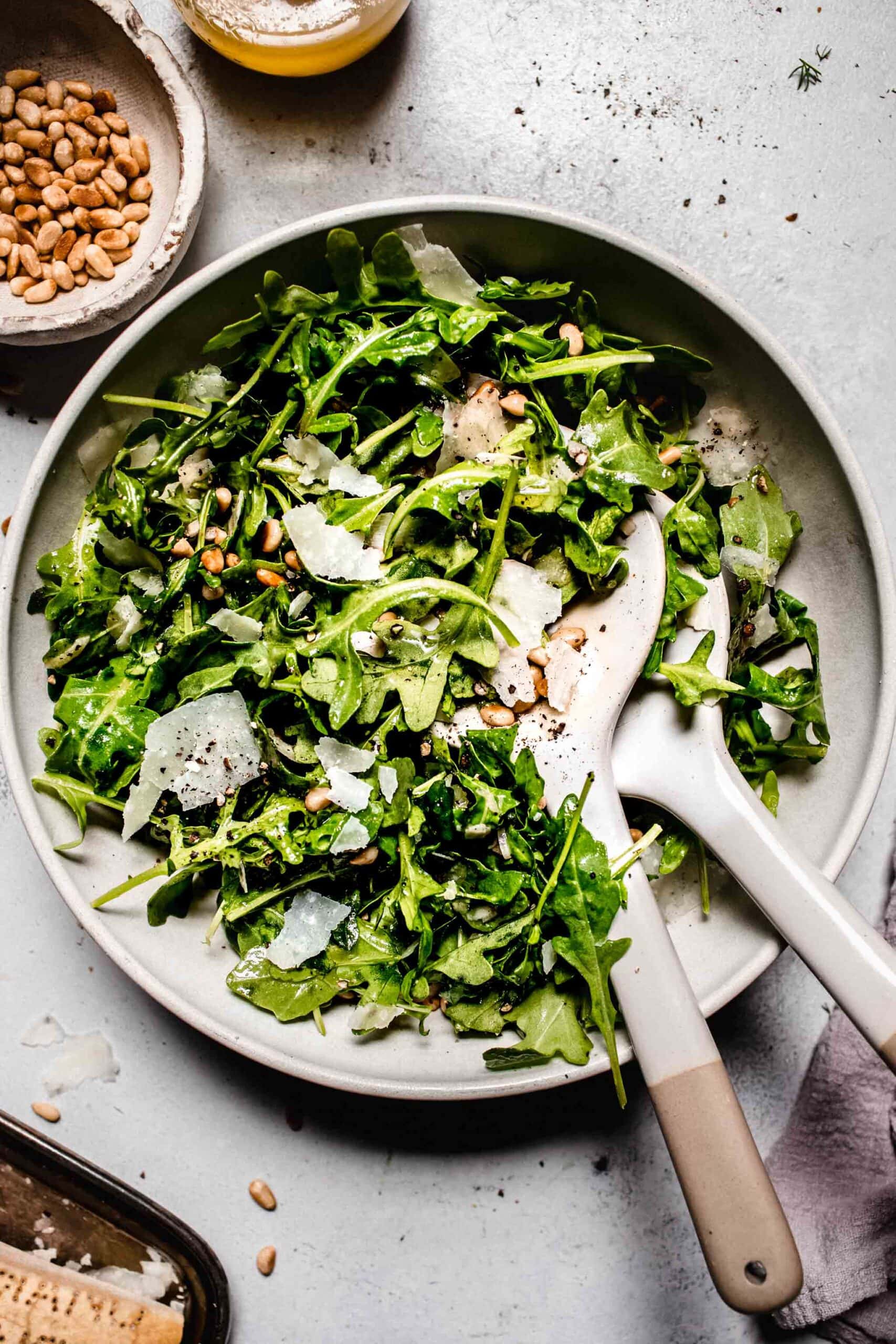 Prepared arugula salad in white bowl with serving spoons.