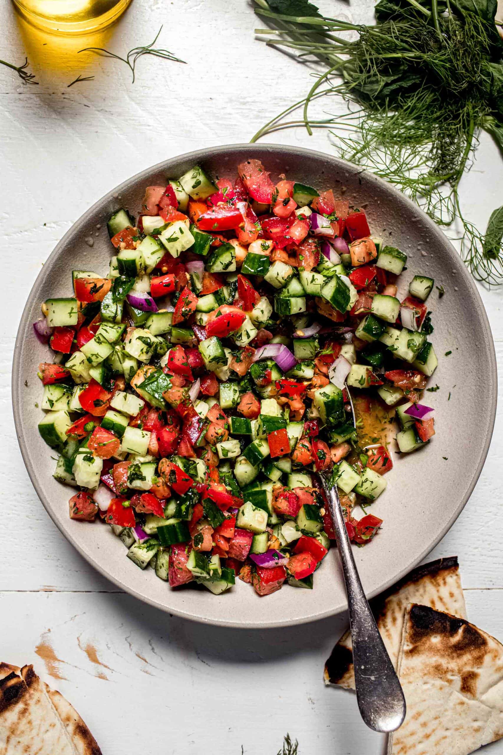 Overhead shot of shirazi salad in white bowl next to pita bread and fresh herbs.