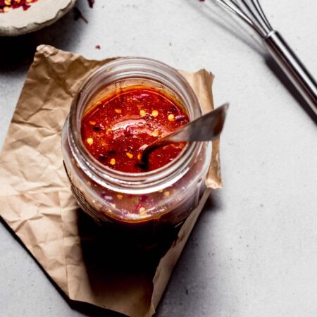 Overhead shot of stir fry sauce in small glass jar next to pepper flakes and whisk.