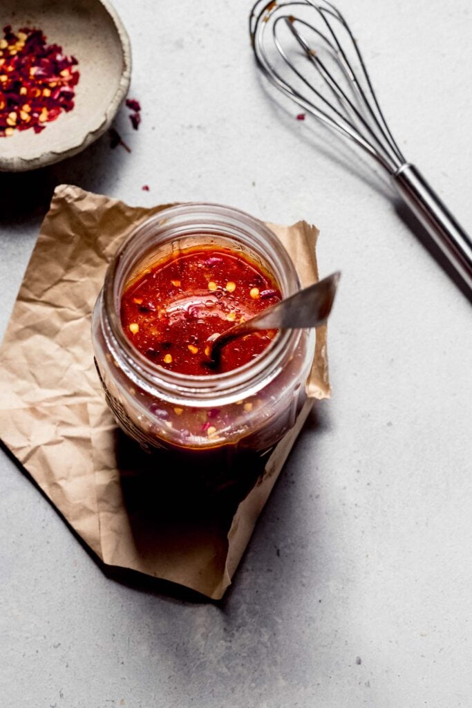 Overhead shot of stir fry sauce in small glass jar next to pepper flakes and whisk.