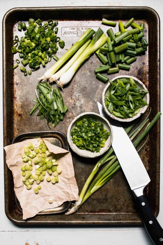 Different cuts of scallions laid out on large baking sheet with knife. 
