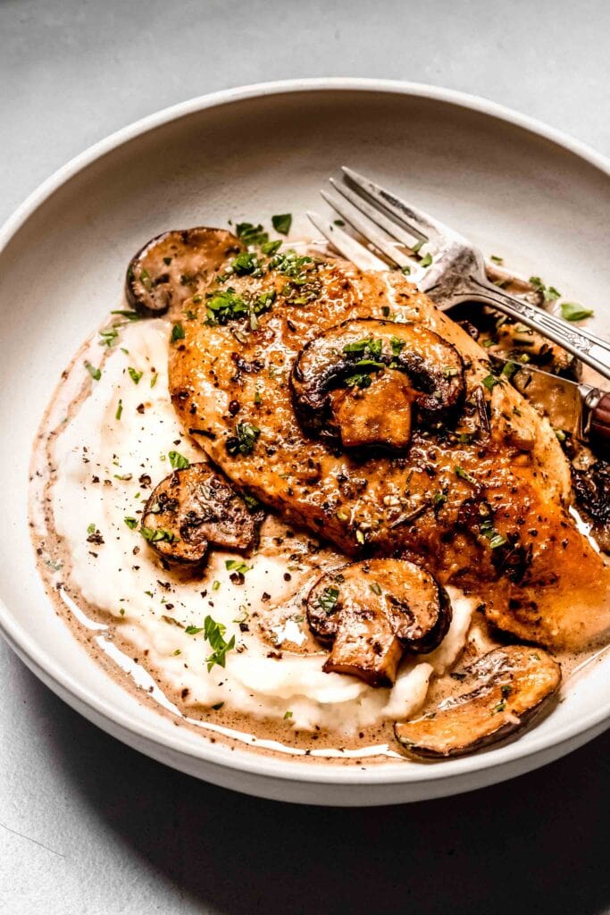 Piece of mushroom chicken in white bowl with mashed potatoes and gravy.