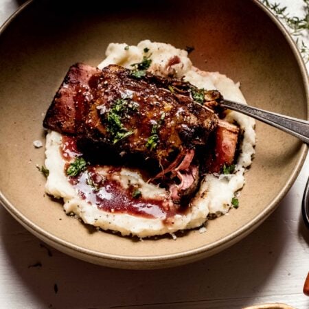 Overhead shot of short rib in brown bowl with mashed potatoes and red wine sauce next to bottle of wine.