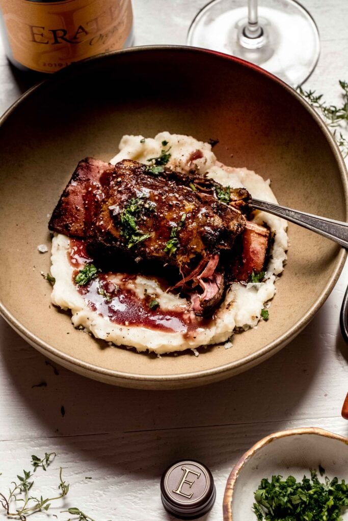 Overhead shot of short rib in brown bowl with mashed potatoes and red wine sauce next to bottle of wine.