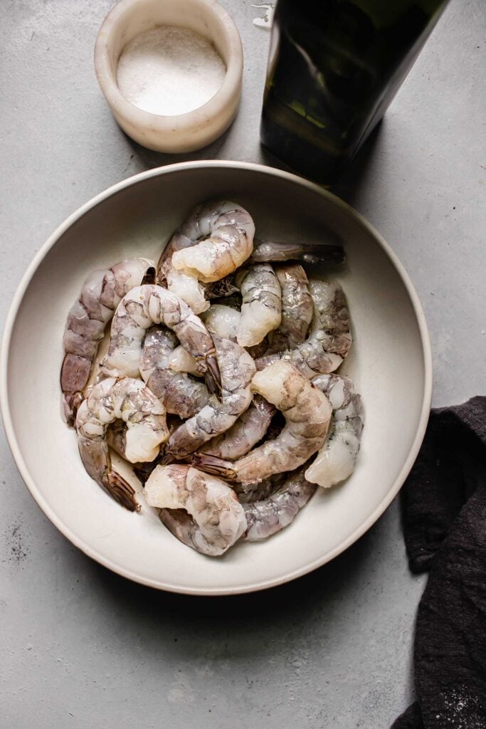 Shrimp in bowl on counter next to bowl of salt and olive oil.