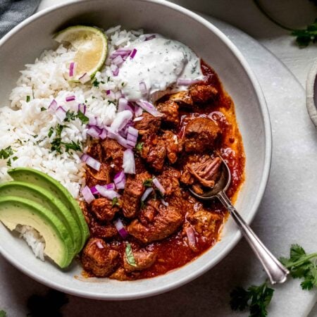 Bowl of mexican lamb stew in white bowl topped with avocado slices and lime wedge.