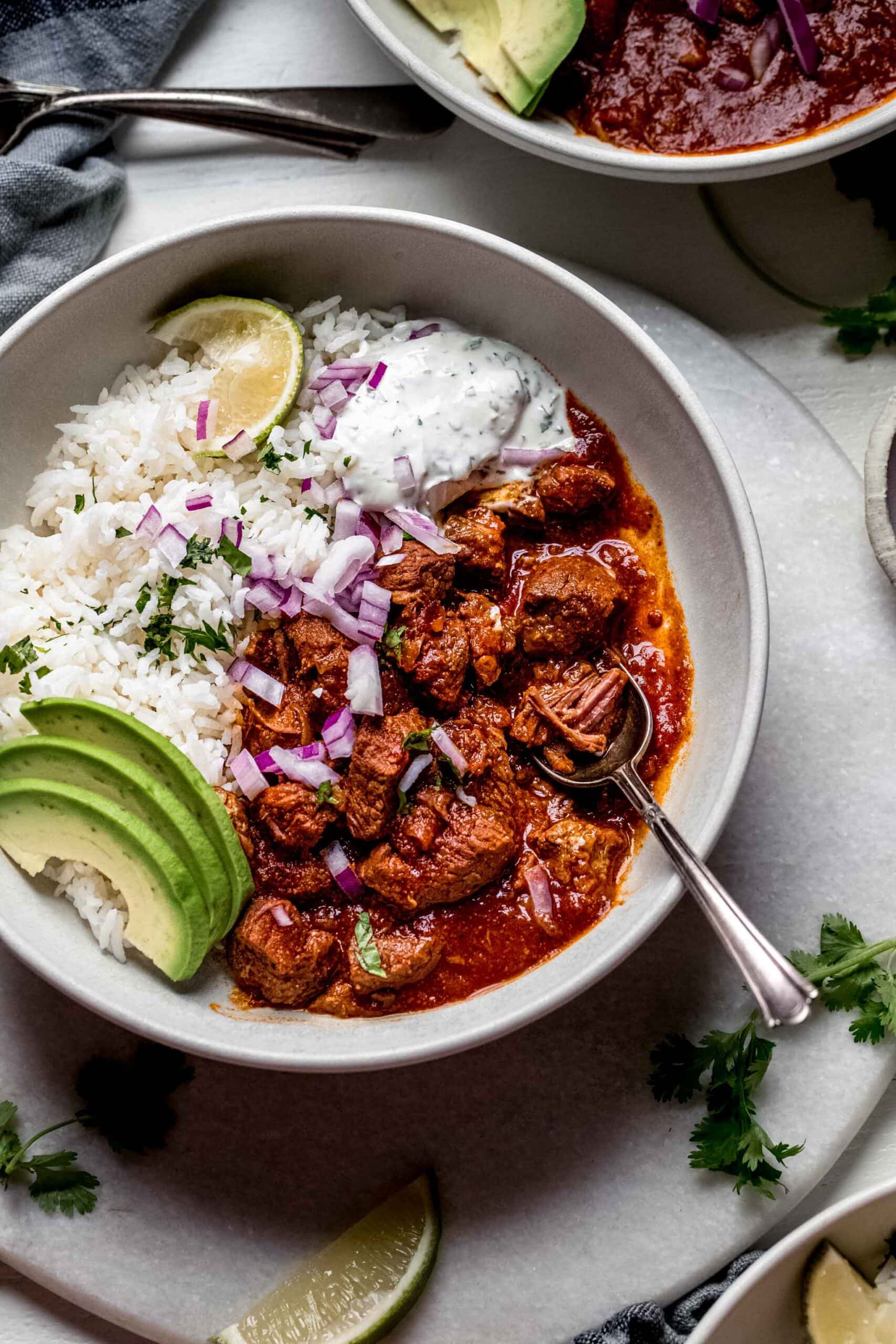 Bowl of mexican lamb stew in white bowl topped with avocado slices and lime wedge.