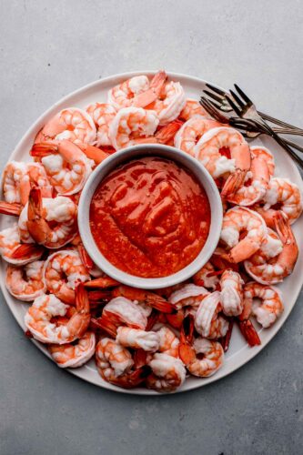 Overhead shot of shrimp arranged on white plate with cocktail forks and bowl of cocktail sauce in the middle.