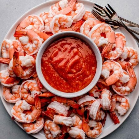 Overhead shot of shrimp arranged on white plate with cocktail forks and bowl of cocktail sauce in the middle.