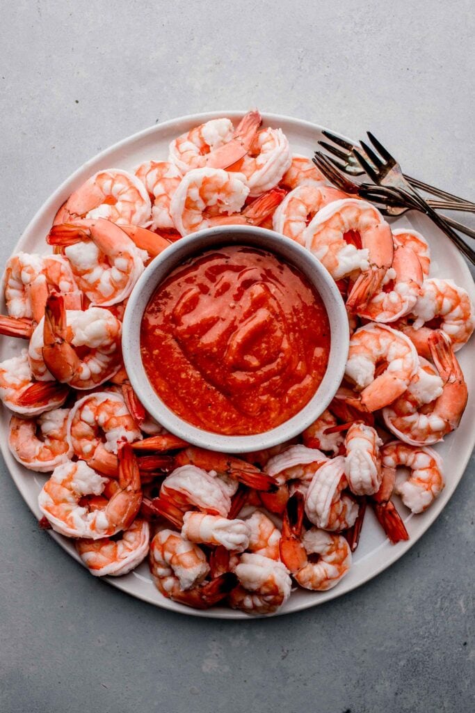 Overhead shot of shrimp arranged on white plate with cocktail forks and bowl of cocktail sauce in the middle.