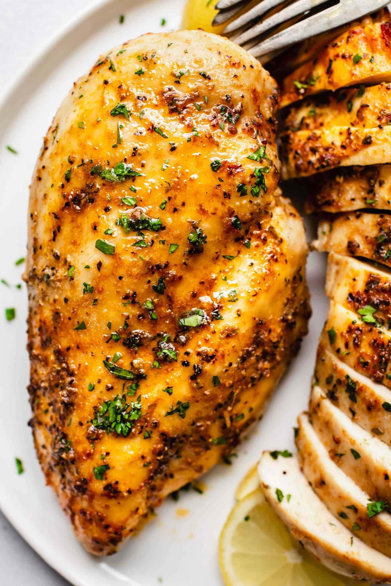 Overhead shot of baked chicken breast on white plate next to lemon slices and sliced chicken breast.