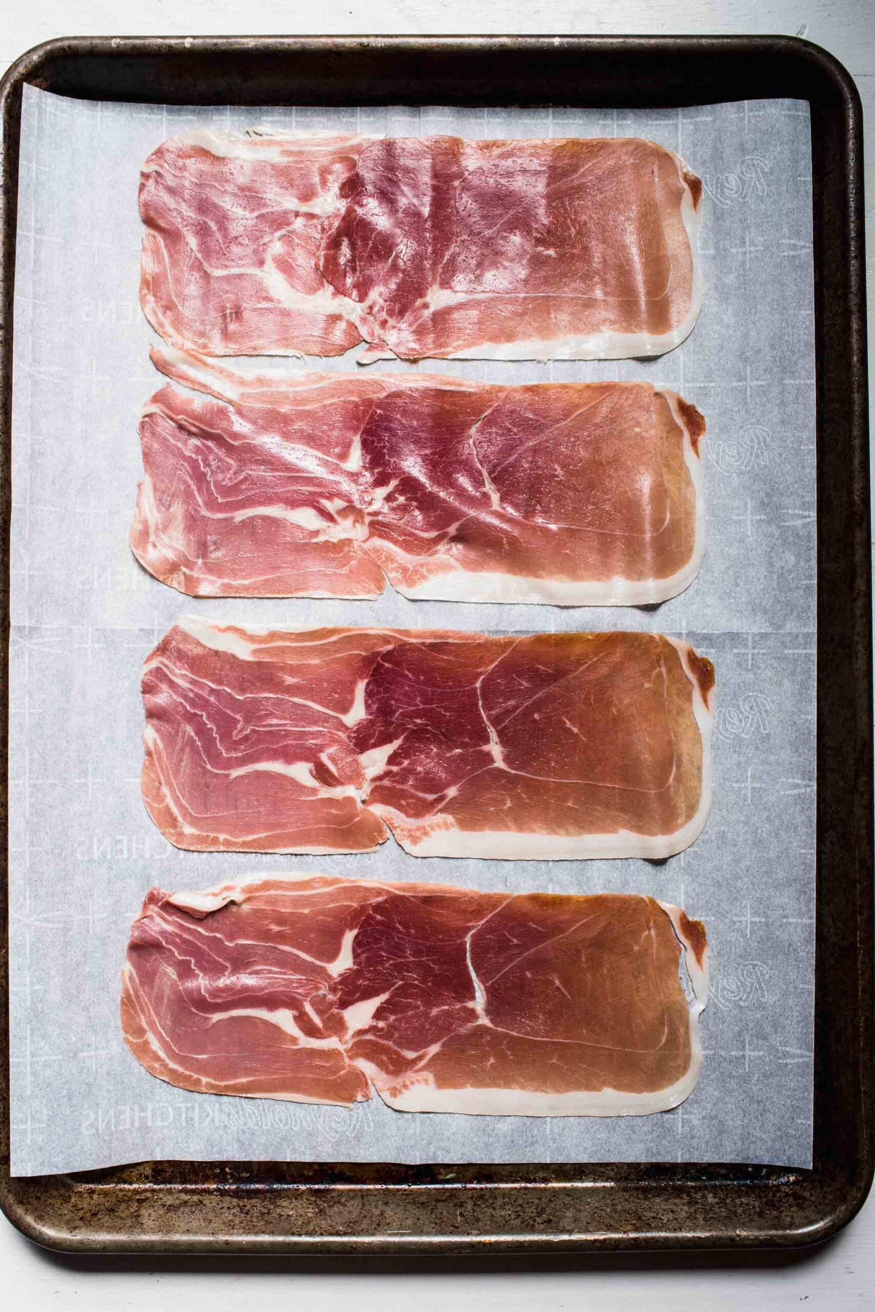 Uncooked prosciutto slices on baking sheet. 