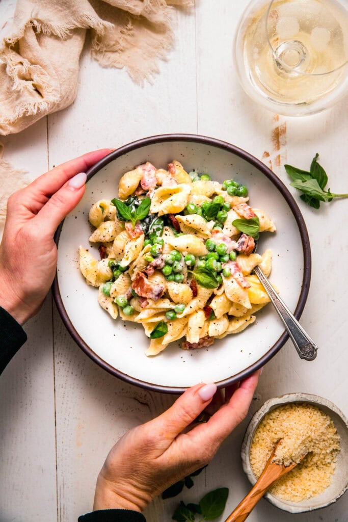 Hands holding bowl of creamy pea and bacon pasta.