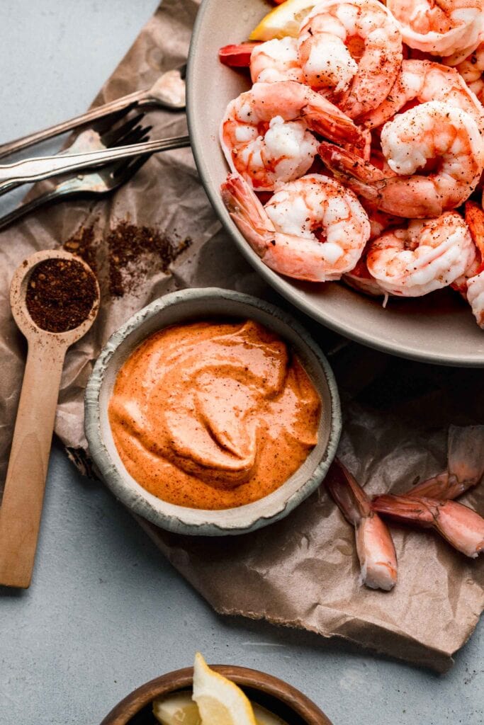 Bowl of remoulade sauce next to small spoon of spices and large bowl of cooked shrimp.