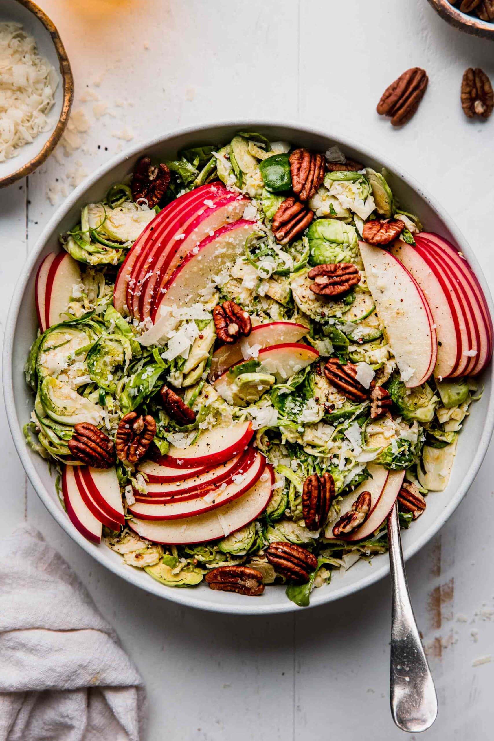 Overhead shot of brussels sporouts & apple salad in white bowl with spoon next to small bowl of parmesan.