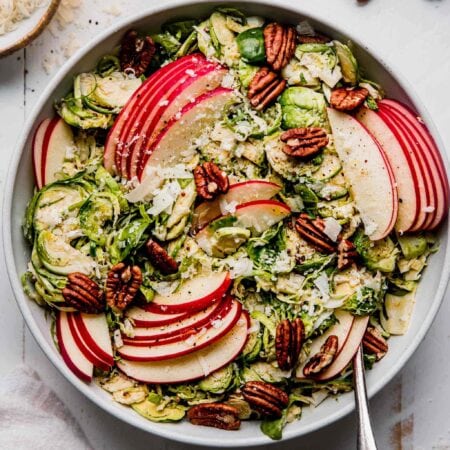 Overhead shot of brussels sporouts & apple salad in white bowl with spoon next to small bowl of parmesan.