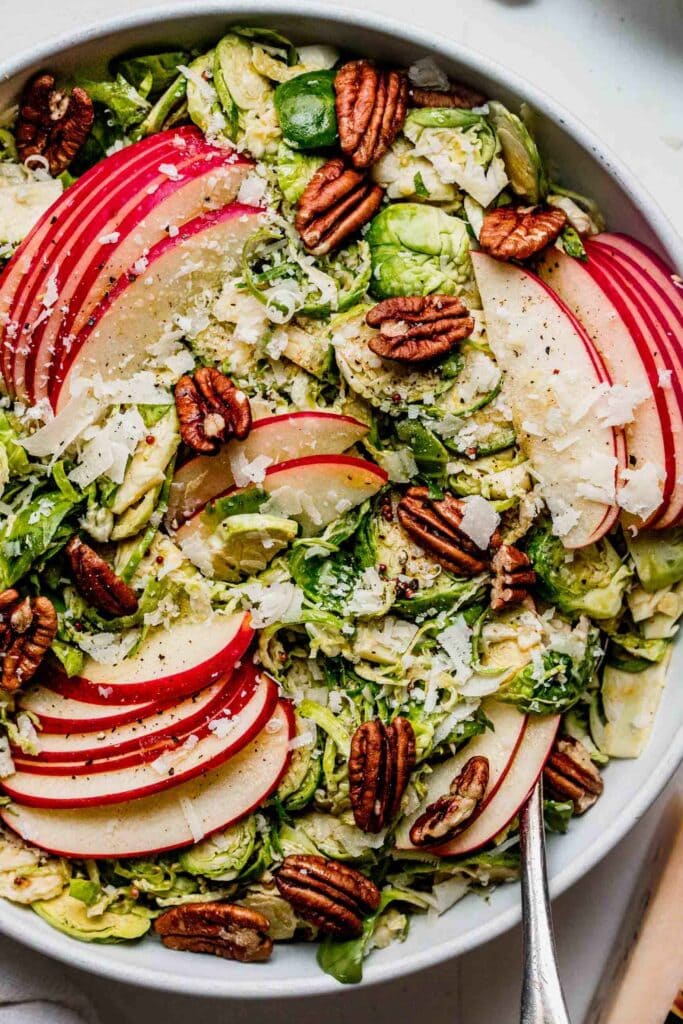 Overhead close up of shaved brussels sporouts & apple salad in white bowl with spoon next to small bowl of parmesan.