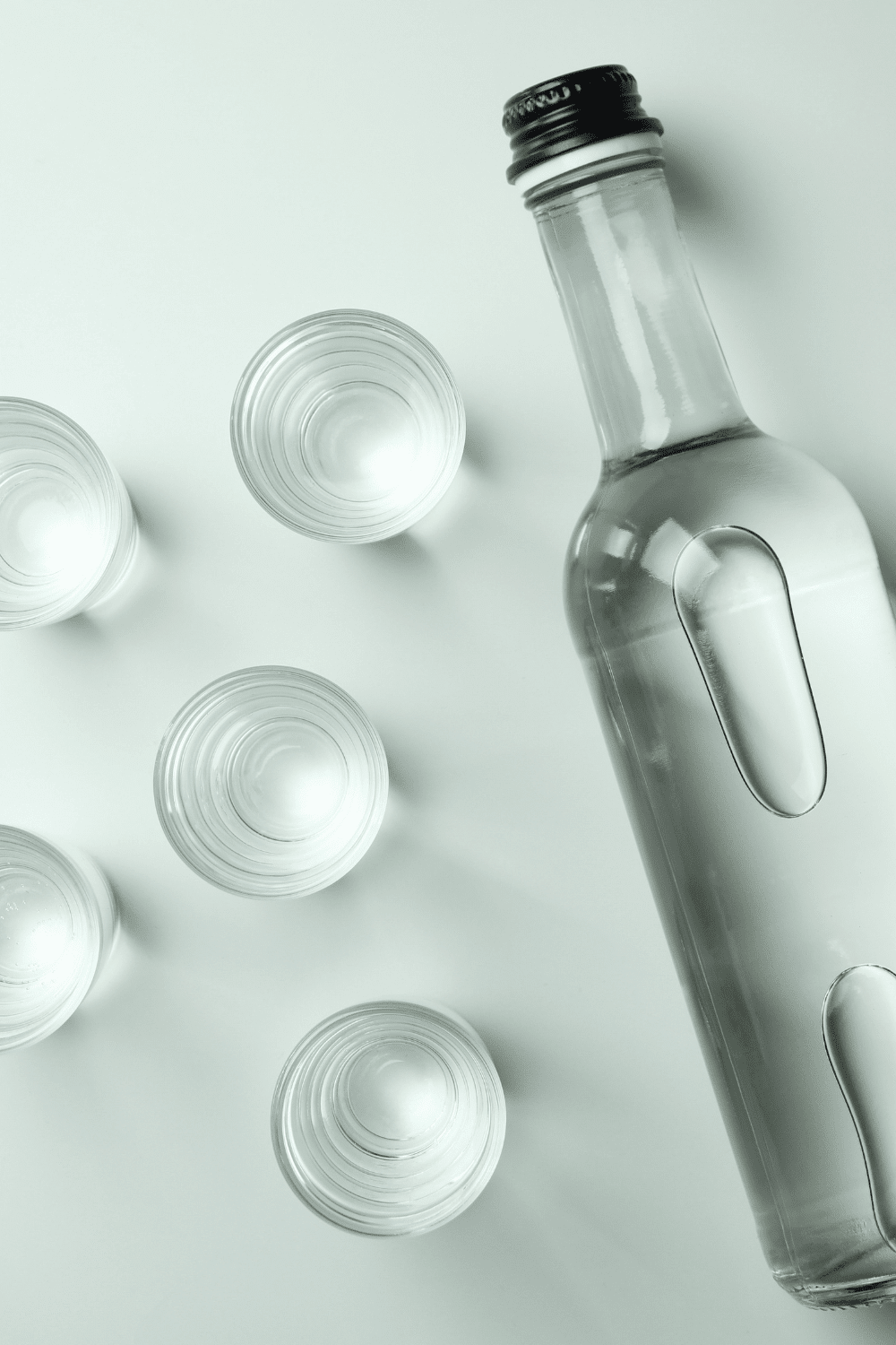Bottle of clear liquor next to shot glasses. 