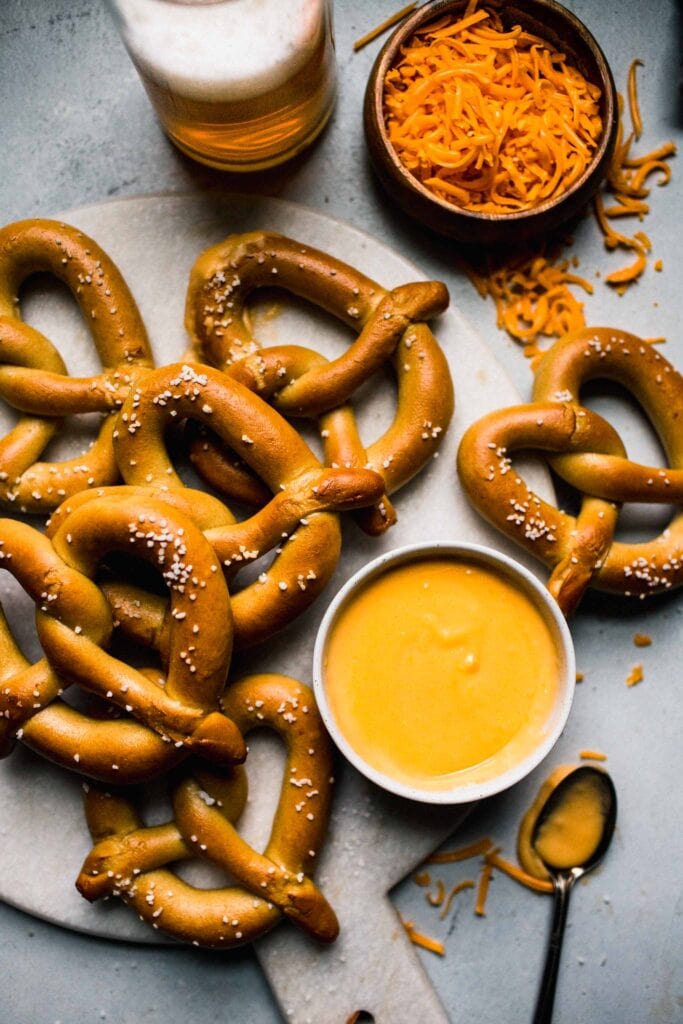 Pretzels on serving platter next to cheese dip, and glass of beer. 