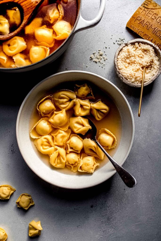 Bowl of tortellini en brodo next to pot of tortellini cooking and small bowl of parmesan. 