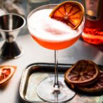 APEROL FIZZ ON SMALL SERVING TRAY GARNISHED WITH DRIED ORANGE WHEEL.