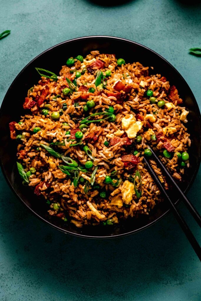 Overhead shot of bacon fried rice in black bowl on teal counter.