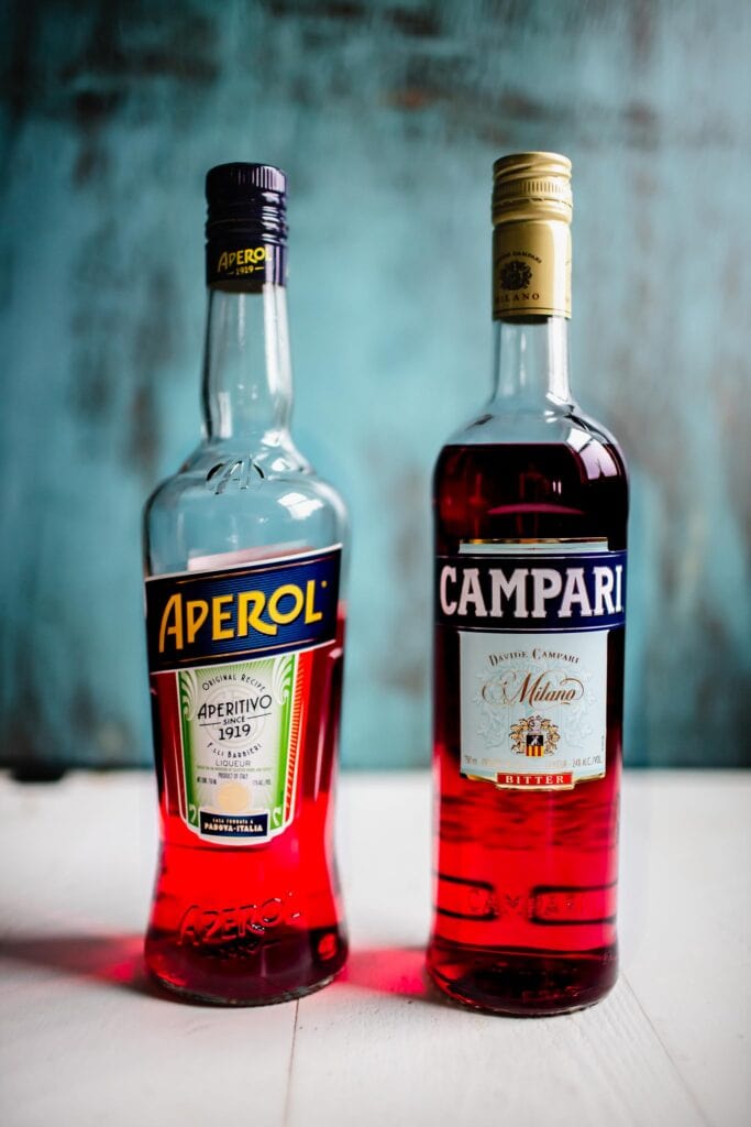 Bottle of campari next to bottle of aperol against blue background.