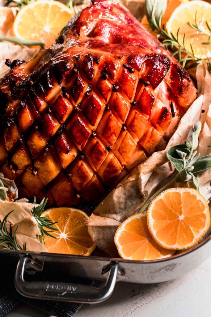 Side vide of ham with glaze in roasting pan surrounded by sliced oranges.