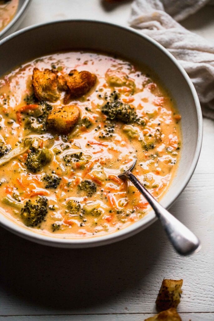 Side view of broccoli cheddar soup in bowl with spoon and toppings. 