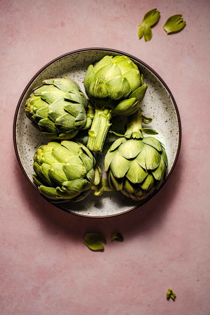 Uncooked artichokes in large speckled bowl on pink counter. 