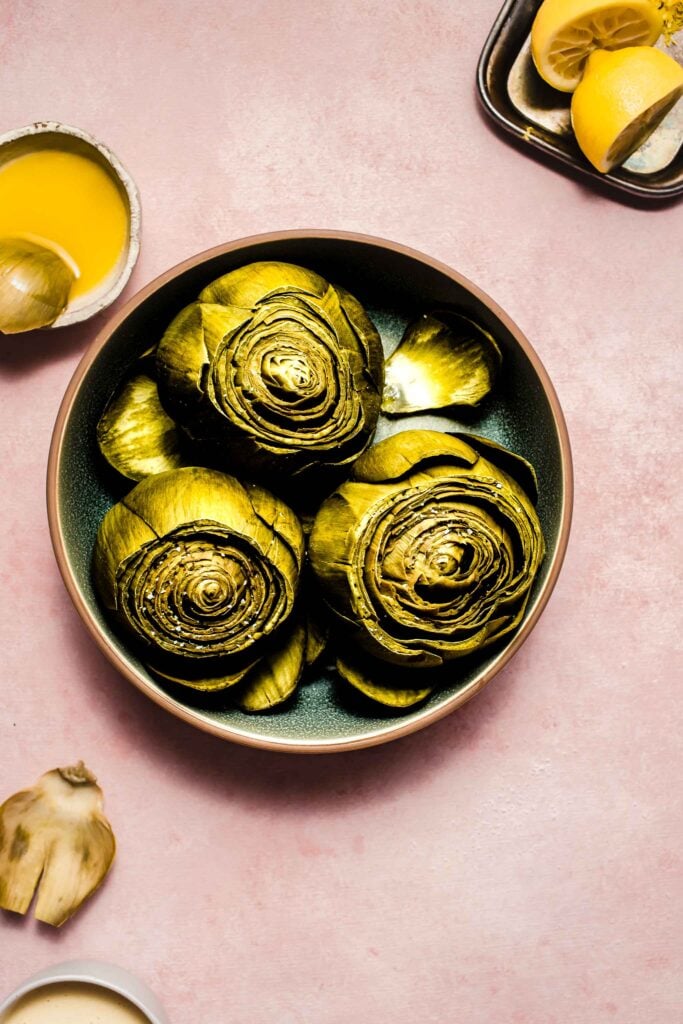 Cooked artichokes in serving bowl next to bowl of melted butter and creamy dipping sauce. 