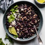 Mexican black beans in grey bowl topped with cotija and lime wedges.