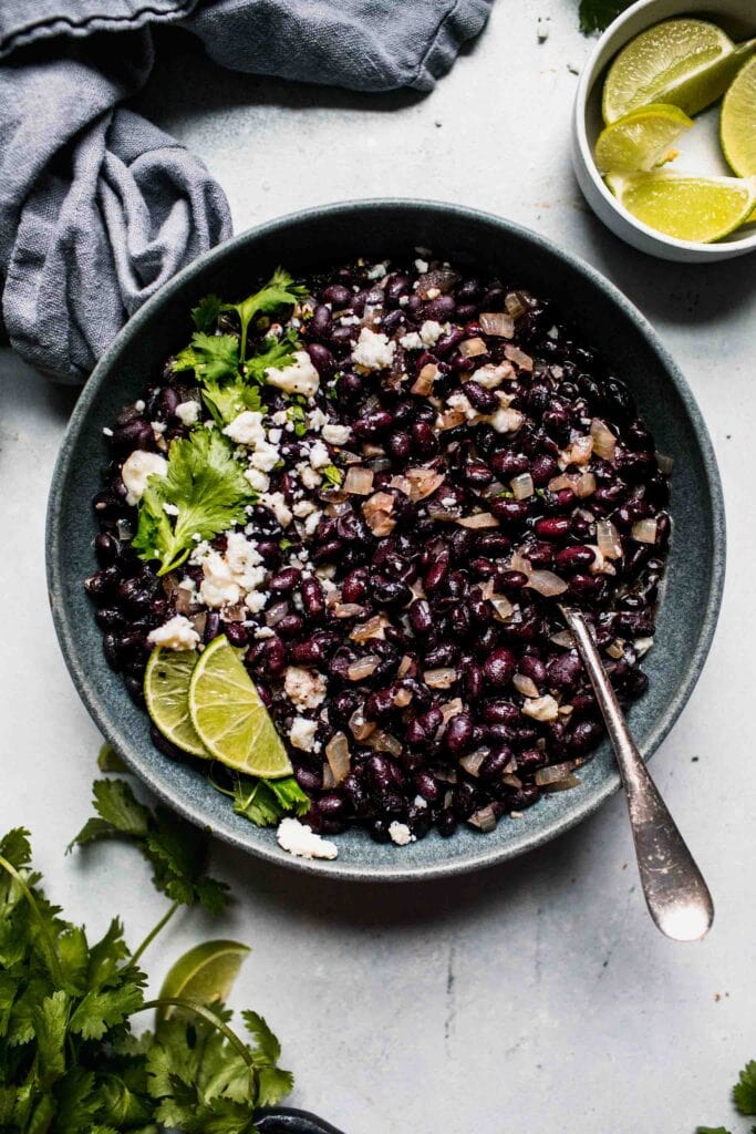 Mexican black beans in grey bowl topped with cotija and lime wedges.