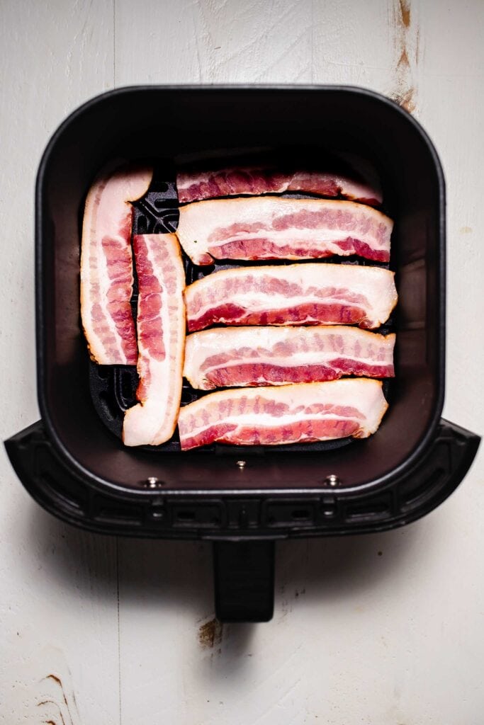 Uncooked bacon slices in air fryer basket. 