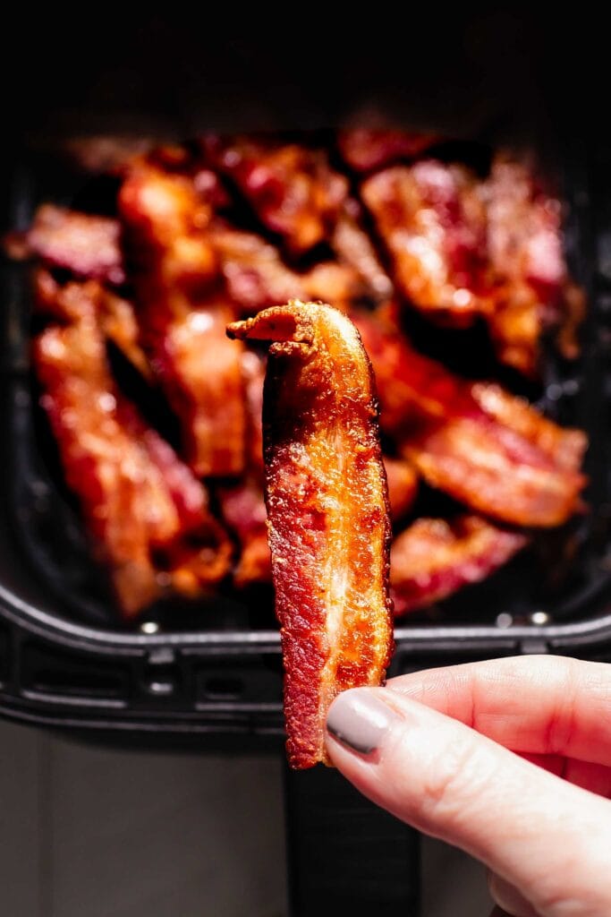 Hand holding strip of crispy bacon over air fryer basket filled with bacon. 