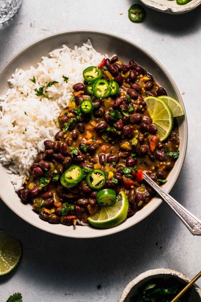 Overhead shot of Black bean curry in white bowl with lime wedges and jalapenos.