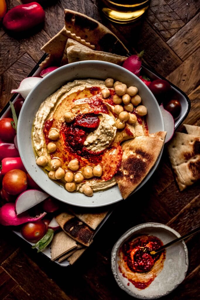 Bowl of harissa hummus on wooden background next to small bowl of hummus paste. 
