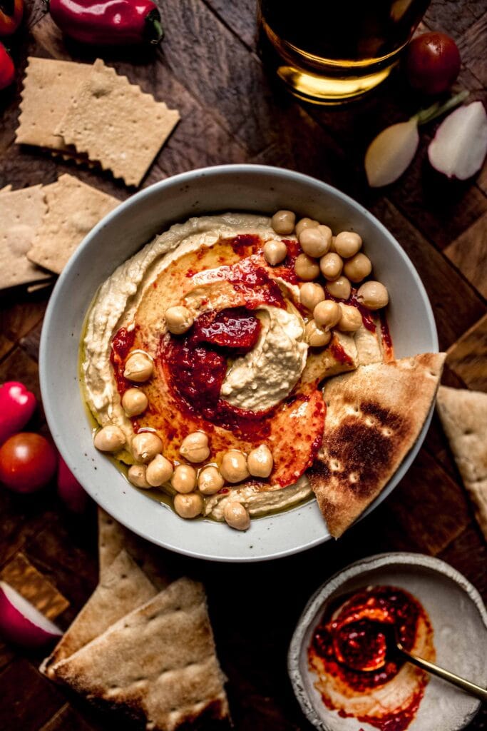 Harissa hummus in bowl surrounded by pita bread and crackers. 