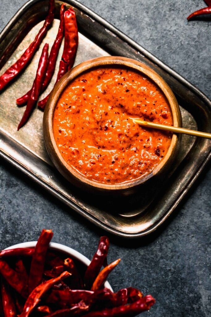 Peri peri sauce in small bowl with spoon next to peppers. 