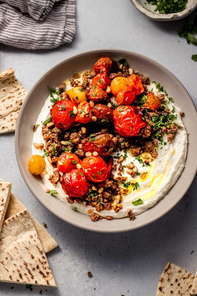 Whipped feta dip in bowl topped with lamb and burst tomatoes, surrounded by pita bread.