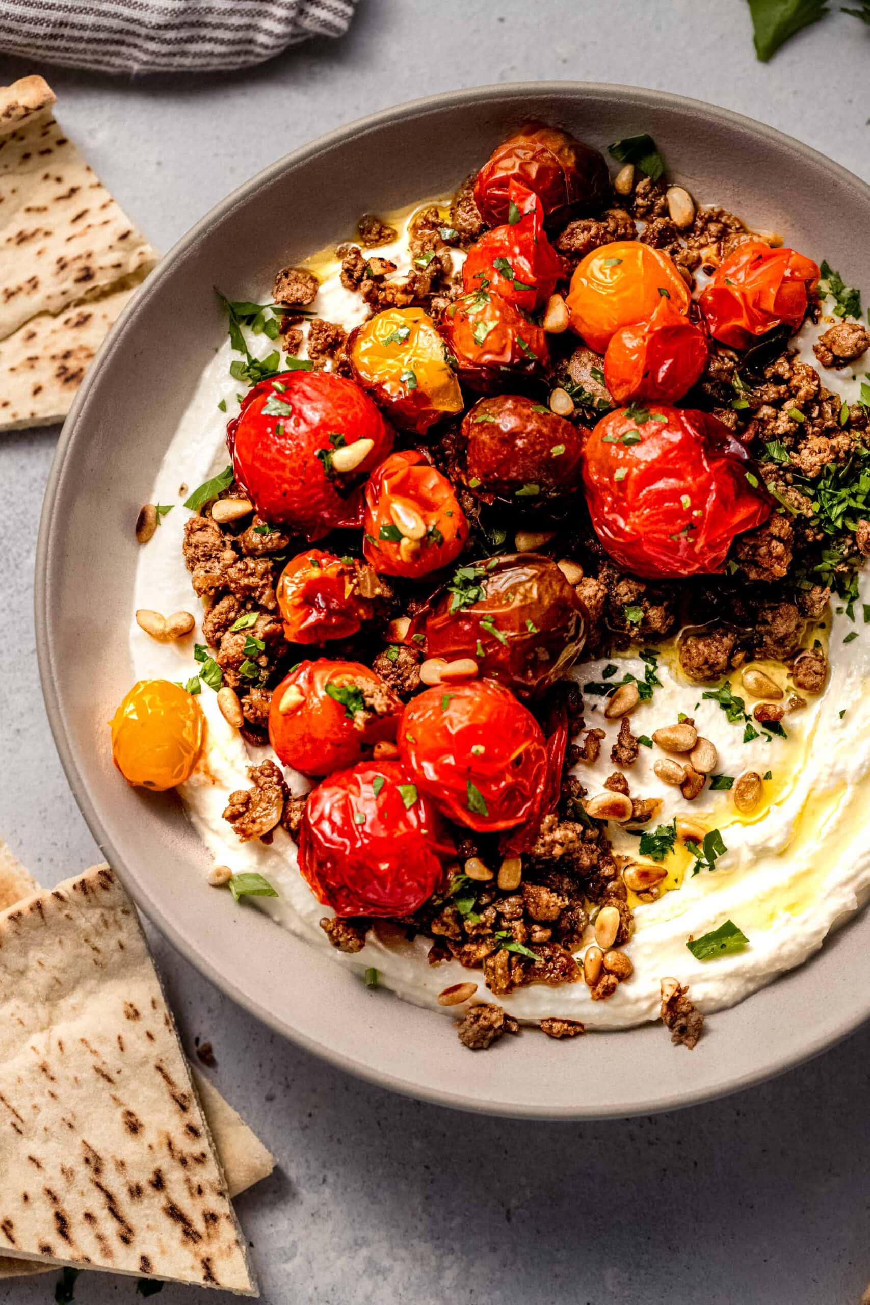 Whipped feta dip in bowl topped with lamb and burst tomatoes, surrounded by pita bread.