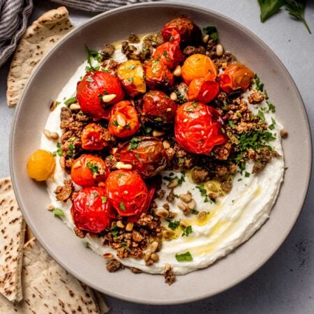 Whipped feta dip in bowl topped with lamb and burst tomatoes, surrounded by pita bread.