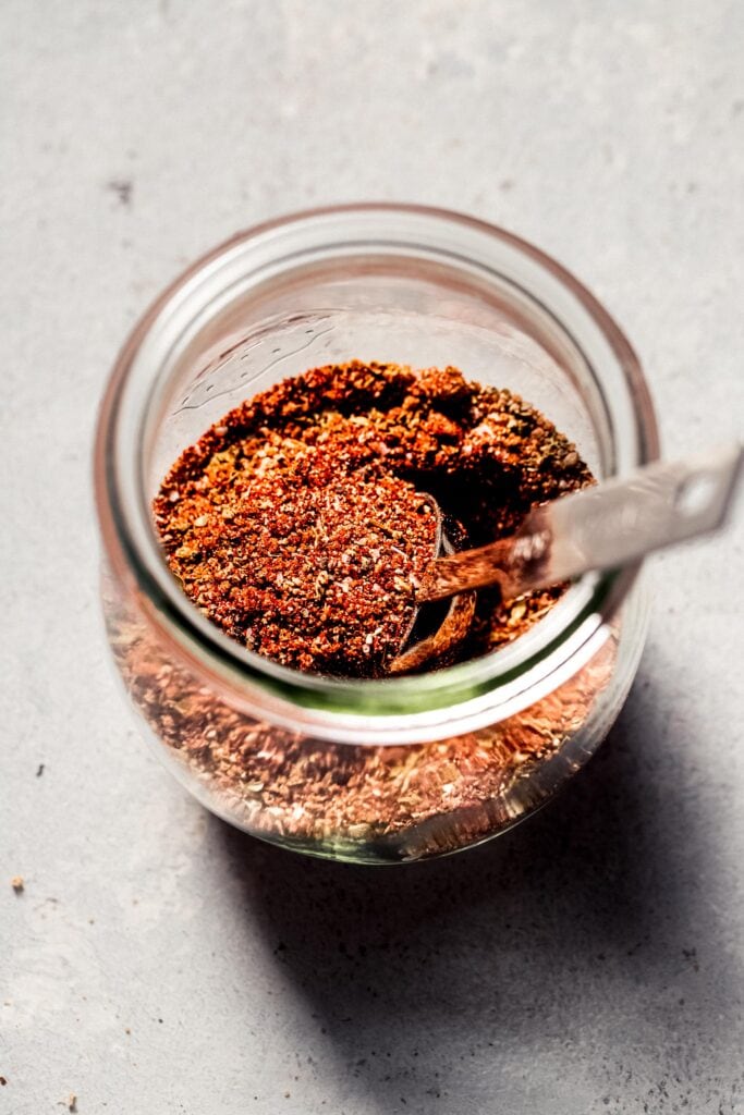 Side view of chicken dry rub in glass jar with spoon.