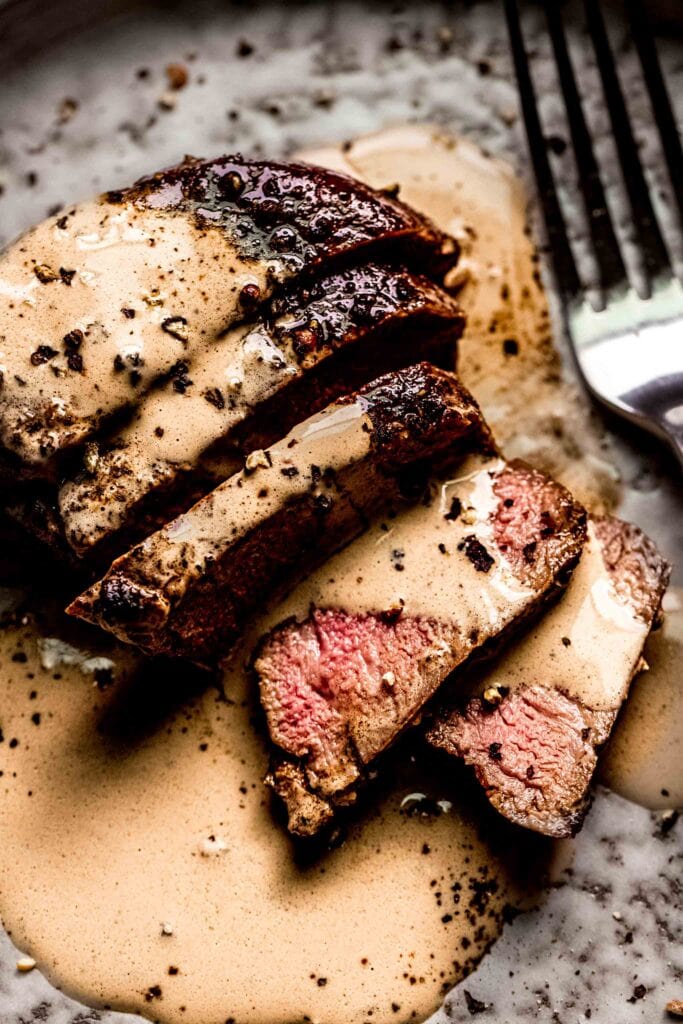 Sliced steak on plate drizzled with peppercorn sauce.