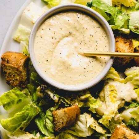 Bowl of vegan caesar dressing on plate of greens with croutons.