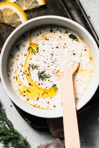 Bowl of yogurt dill sauce with spoon and slices of lemon, drizzled with olive oil.